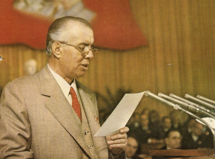 Enver Hoxha í ræðustól með fána albanska kommúnistaflokksins í baksýn.